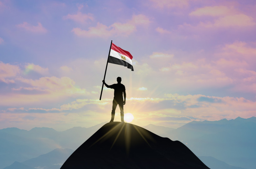 Ägypten - Sharm el Sheikh Eine Silhouette eines Mannes steht auf einem Berggipfel und hält die ägyptische Flagge. Die Sonne geht im Hintergrund auf, der Himmel ist rosa-blau gefärbt.