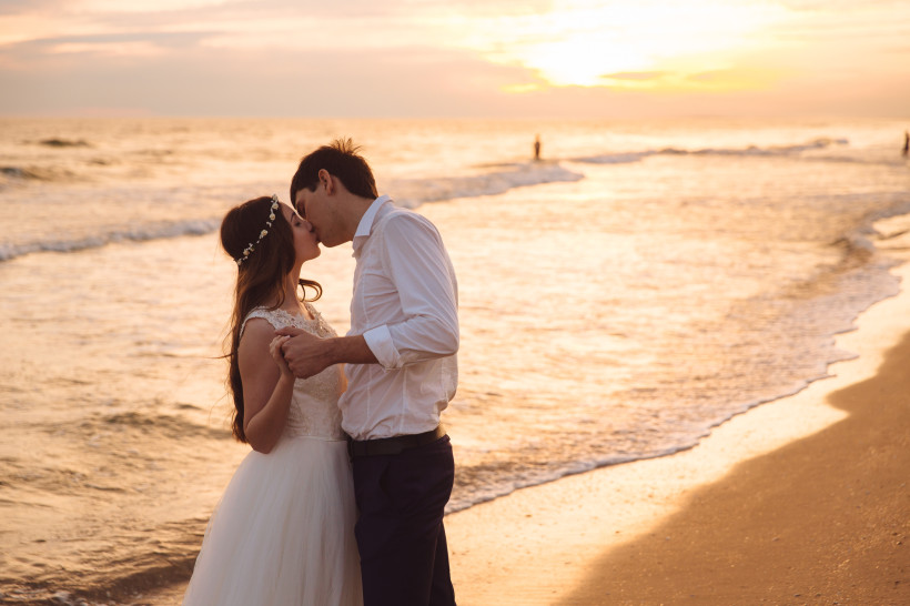 Romantischer Sonnenuntergang – unvergessliche Hochzeitsreise für Paare Frisch verheiratetes Paar küsst sich bei Sonnenuntergang am Strand – perfekte Kulisse für eine unvergessliche Hochzeitsreise