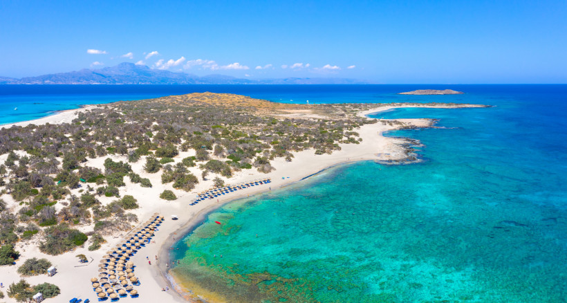 Luftaufnahme von Chrissi Island mit Sandstrand, flachem türkisblauem Meer und Sonnenschirmen entlang der Küste