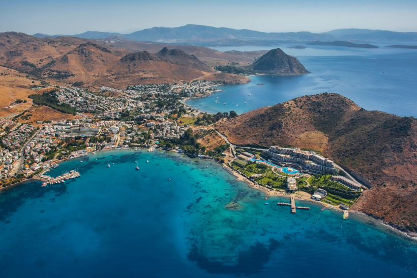 Luftaufnahme einer türkisblauen Bucht mit Hotelanlage, Yachthafen und bergiger Küstenlandschaft
