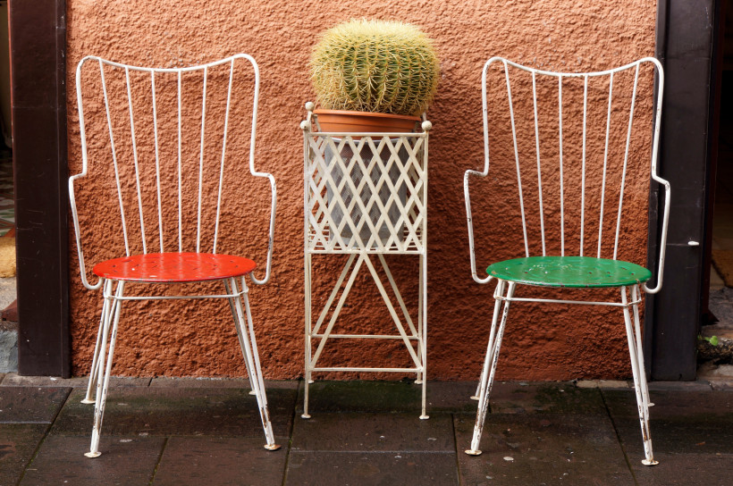 Bunte Stühle mit Kaktus – rustikaler Terrassenstil auf den Kanarischen Inseln Zwei Metallstühle mit roten und grünen Sitzen und ein Kaktus in einem Topf auf einem Eisengestell vor einer Terrakottawand
