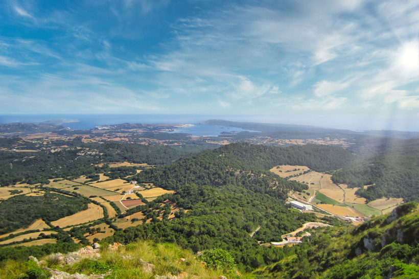 Weitläufiger Panoramablick über die hügelige Landschaft von Menorca. In der Ferne sind Felder, Wälder und die Küstenlinie mit dem Meer zu sehen. Die Szenerie ist von leicht bewölktem, sonnigem Himmel überzogen, mit weichem Licht von der rechten Seite.