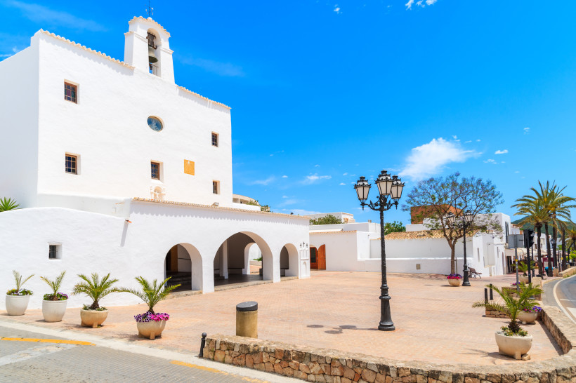 Ibiza - Sant Josep de sa Talaia Weiße Kirche mit Arkaden und Glockenturm im Ort Sant Josep de sa Talaia auf Ibiza. Vor dem Gebäude befindet sich ein gepflasterter Platz mit Blumenkübeln, Laternen und Palmen unter blauem Himmel.