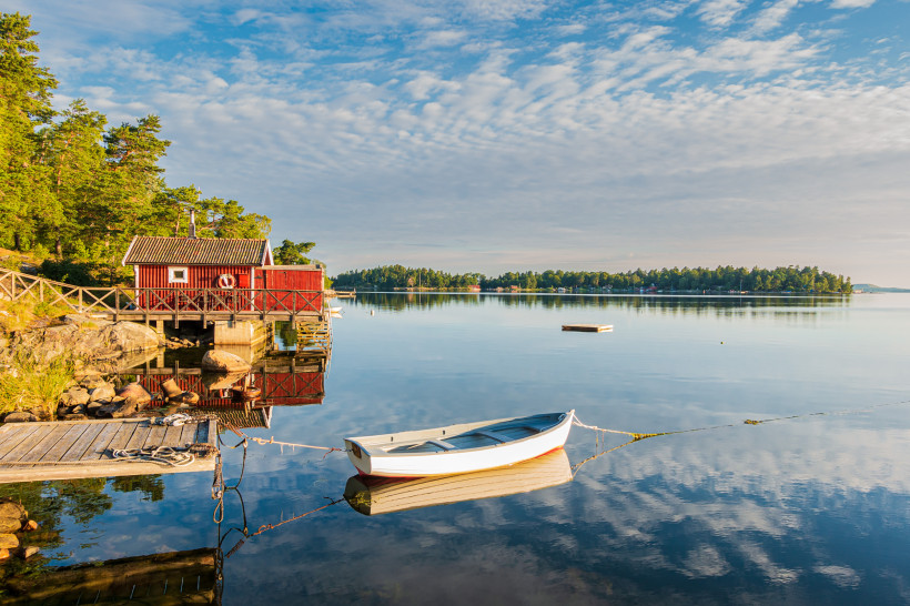 Schwedischer Schärengarten vor Stockholm mit Boot und roten Holzhäusern am Wasser Rote Holzhäuser am Ufer und kleines Ruderboot auf ruhigem Wasser im Schärengarten vor Stockholm, Schweden