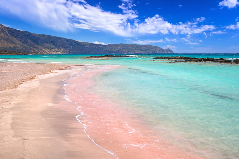 Küstenansicht am Elafonissi-Strand mit Lagune und Bergen unter blauem Himmel Elafonissi-Strand mit hellem Sand, türkisfarbenem flachem Wasser und Bergkette im Hintergrund