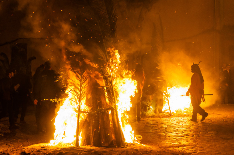 Mallorca Nachtaufnahme eines traditionellen Festes mit mehreren brennenden Holzstapeln. Eine Person in einem roten Kostüm mit Hörnern, die einen Teufel darstellen, steht neben den Flammen. Weitere Menschen in dunkler Kleidung beobachten das Geschehen im Hintergrun