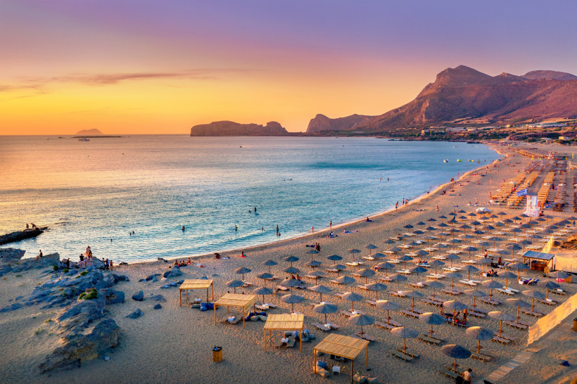 Kreta Ein langer Sandstrand auf Kreta bei Sonnenuntergang mit zahlreichen Sonnenliegen und Schirmen. Die Küste ist von sanften Wellen umspült, während die Sonne den Himmel in warmen Farben von Gelb bis Violett taucht. Im Hintergrund ragen Bergketten und vorgela