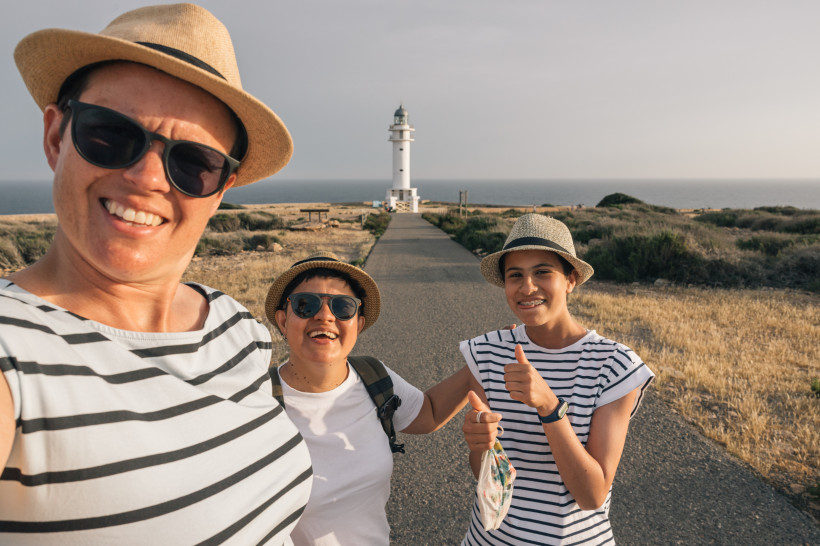 Formentera Drei Frauen machen ein Selfie auf einem sonnigen Küstenweg, im Hintergrund ein weißer Leuchtturm am Ende des Weges. Alle tragen Hüte, Sonnenbrillen und haben sommerliche Kleidung an. Die Stimmung ist fröhlich und ausgelassen, eine der Frauen zeigt einen D