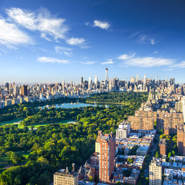 New York Panoramaaufnahme von New York City mit dem Central Park im Vordergrund. Der grüne Park bildet einen starken Kontrast zur umliegenden Skyline aus hohen, modernen Gebäuden. Der Himmel ist blau mit wenigen Wolken.