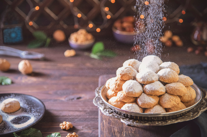 Ägypten - Marsa Alam Ein kunstvoll arrangierter Haufen orientalischer Kekse, die großzügig mit Puderzucker bestreut werden. Im Hintergrund liegen Nüsse und Minzblätter auf einem rustikalen Holztisch.