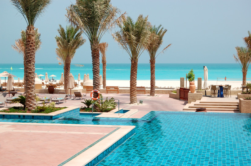 Poolanlage mit Palmen und Liegen am Strand in Abu Dhabi, im Hintergrund das Meer