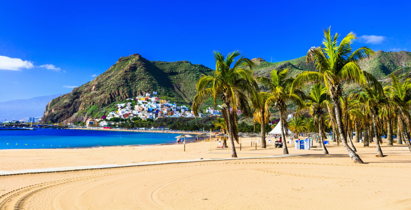Las Teresitas in der Nähe von Santa Cruz. Kanarische Inseln Sandstrand mit Palmen vor einem türkisblauen Meer, im Hintergrund bunte Häuser am Berghang unter klarem, blauem Himmel