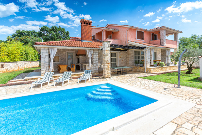 Kroatien Großes Ferienhaus mit rosa Fassade, Natursteinmauern und rotem Ziegeldach. Im Vordergrund befindet sich ein Pool mit Liegestühlen. Der Garten ist gepflegt mit Rasen, Olivenbaum und überdachter Terrasse mit Grill.