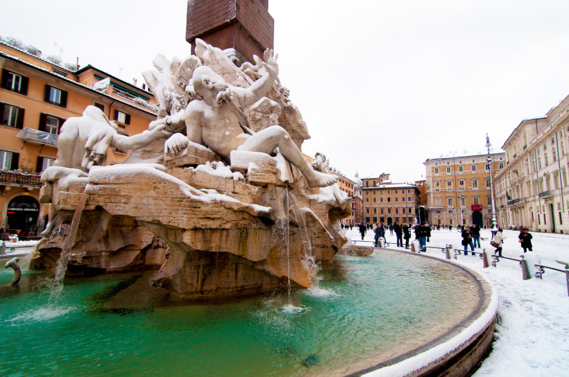 Rom Verschneiter Vierströmebrunnen auf der Piazza Navona in Rom. Die barocke Brunnenfigur ist mit einer Schneeschicht bedeckt, Wasser fließt weiterhin in das grünlich schimmernde Becken. Menschen spazieren durch das winterliche Stadtzentrum.