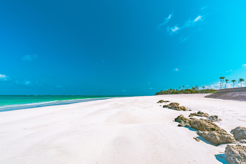 Kenia Ein weiter, menschenleerer Sandstrand mit strahlend weißem Sand und türkisblauem Meer unter wolkenlosem Himmel. Auf der rechten Seite Palmen und einige Felsen am Strand, im Hintergrund tropische Vegetation