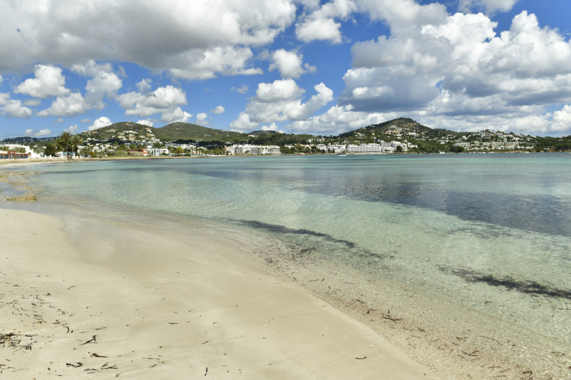 Talamanca Strand mit hellem Sand und ruhigem, türkisfarbenem Wasser, im Hintergrund Hügel mit Bebauung unter wolkigem Himmel