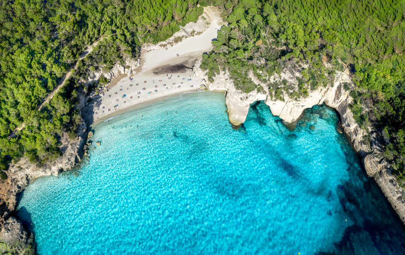 Menorca - Cala Mitjana Luftaufnahme Cala Mitjana und Mitjaneta auf Menorca: feiner Sandstrand, umgeben von grünen Pinienwäldern, mit türkisblauem Wasser, in dem einige Badegäste schwimmen.