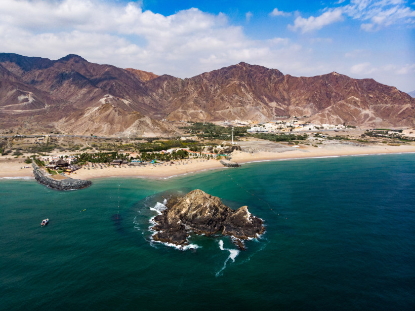Fujairah, Luxusferien  Luftaufnahme eines Strandes mit Bergen im Hintergrund und einer kleinen Felseninsel im Meer
