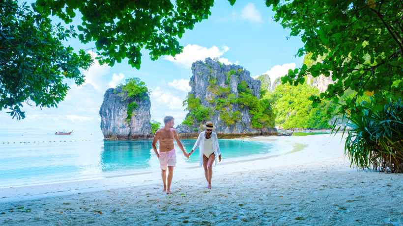 Thailand Das Bild zeigt ein Paar, das barfuß Hand in Hand an einem tropischen Sandstrand entlangspaziert. Im Hintergrund liegt kristallklares, türkisfarbenes Wasser, das sanft an den Strand schwappt. Die Szene wird eingerahmt von dichtem, grünem Blattwerk und mark