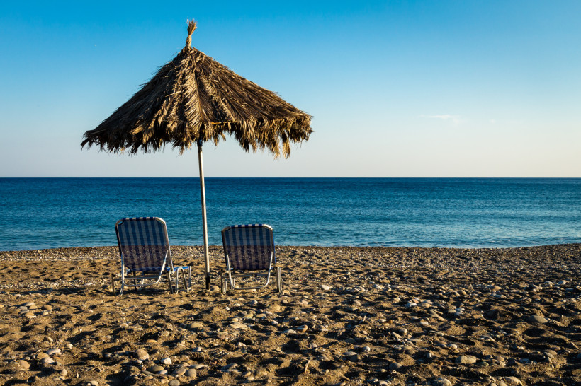 Zwei Liegestühle unter einem Strohdach am ruhigen Kiesstrand auf Kreta mit Blick aufs Meer