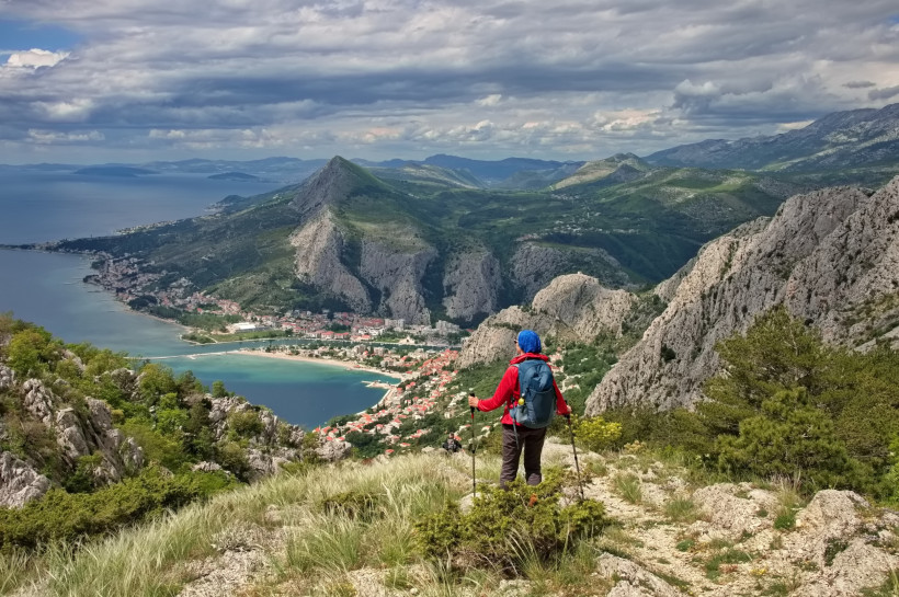 Kroatien Wanderer mit roter Jacke und Rucksack steht auf einem Bergpfad mit Blick auf eine Küstenstadt zwischen Meer und hohen, felsigen Bergen. Der Himmel ist bewölkt, das Panorama weitläufig.