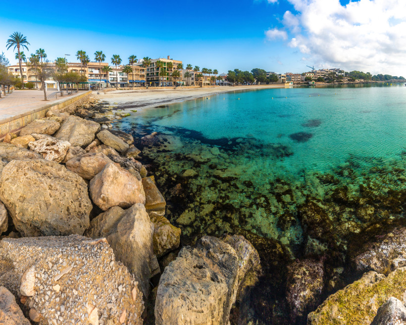 Küstenpromenade und Bucht in Colonia de Sant Jordi mit Felsen im Vordergrund und türkisfarbenem Meer
