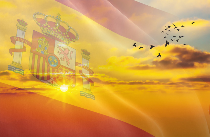 Transparente spanische Flagge mit Wappen weht über einem stimmungsvollen Sonnenuntergang. Der Himmel ist orange-gold gefärbt, einige Wolken sind zu sehen. Rechts fliegt ein Vogelschwarm in Formation.
