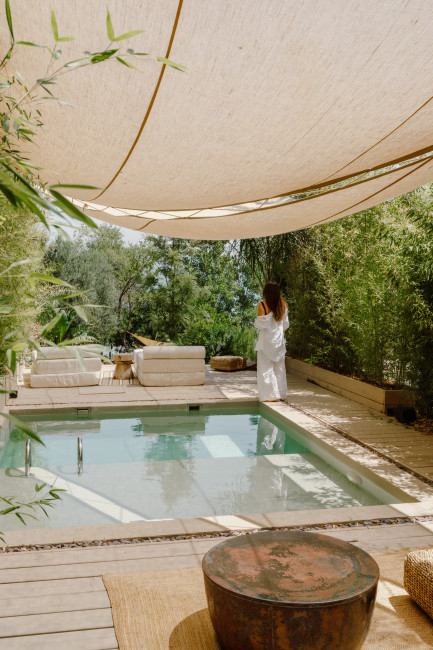 Eine Frau im weißen Bademantel steht auf einer Holzterrasse neben einem kleinen Pool, umgeben von Bambus und mediterraner Vegetation. Über dem Bereich spenden helle Stoffbahnen Schatten, die Atmosphäre ist ruhig und entspannt