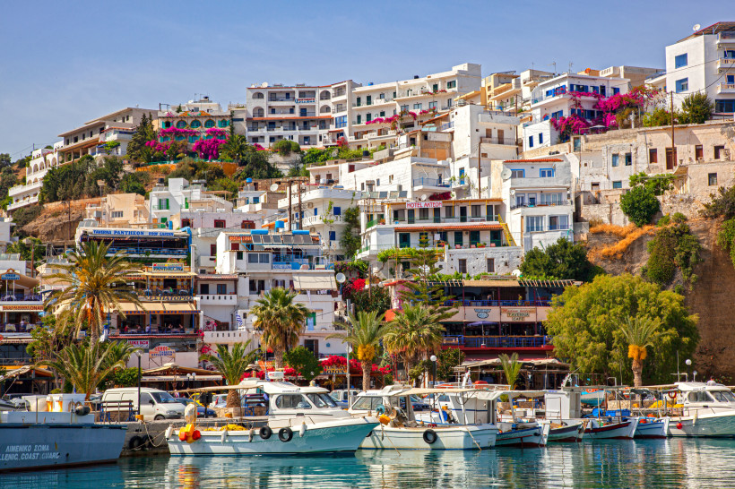 Lebendige Hafenstadt mit weißen und farbigen Häusern an einem Hügel, Palmen, blühenden Pflanzen und zahlreichen Fischerbooten und Yachten im klaren Wasser