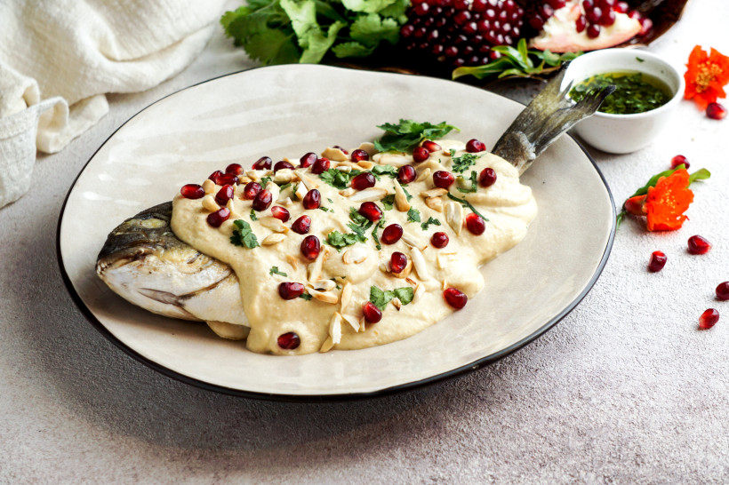 Ägypten - Hurghada Ein ganzer Fisch auf einem Teller, überzogen mit cremiger Tahini-Sauce, garniert mit Granatapfelkernen, Mandelsplittern und frischen Kräutern. Im Hintergrund Granatapfel, frische Kräuter, eine grüne Sauce und eine orange Blüte.