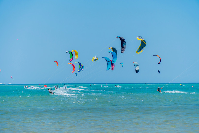 Ägypten - El Gouna Farbenfrohe Kites segeln am blauen Himmel über dem türkisfarbenen Meer. Zahlreiche Kitesurfer gleiten dynamisch über das Wasser, erzeugen Gischt und nutzen den kräftigen Wind. Die Szene vermittelt Energie, Freiheit und Sportbegeisterung an einem sonnigen