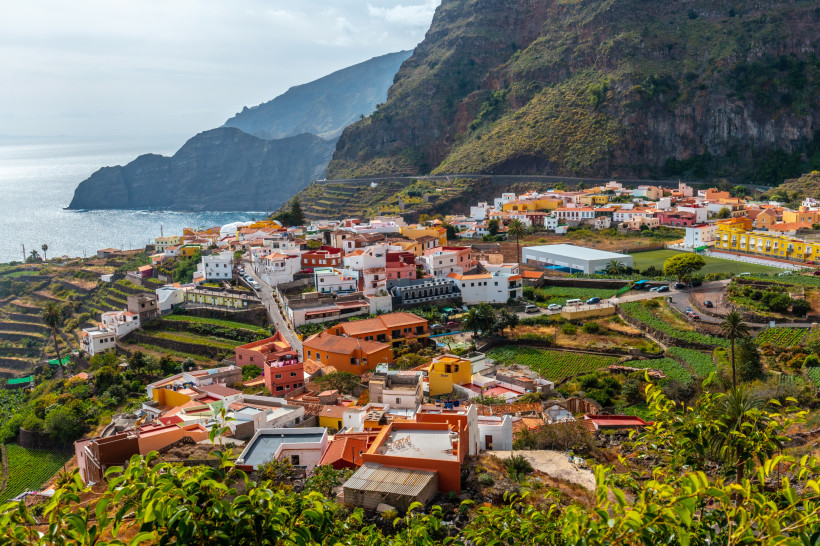 Blick auf ein Küstendorf auf La Gomera mit bunten Häusern, Terrassenfeldern und steilen Bergen am Meer