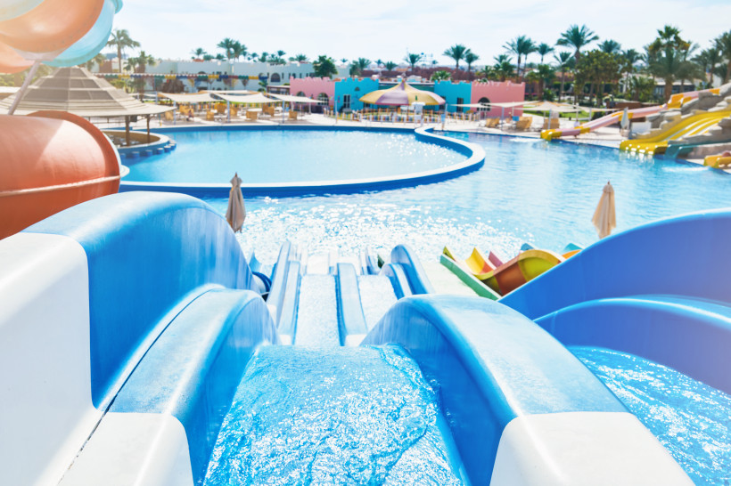 Ägypten - Makadi Bay Blick von oben auf mehrere blaue Wasserrutschen, die in einen großen, sonnigen Pool führen. Rund um das Becken stehen Sonnenliegen und bunte Sonnenschirme. Im Hintergrund sind weitere Rutschen in Gelb, Orange und Rot sowie Palmen und bunte Gebäude zu sehe