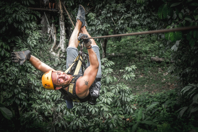 Thailand - Phuket Ein junger Mann mit Helm und Klettergurt schwingt lachend an einem Zipline-Seil durch einen dichten, grünen Dschungel. Er trägt eine Kamera an seinem Gurt und hebt fröhlich den Daumen. Im Hintergrund sind viele tropische Pflanzen zu sehen.