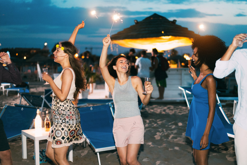 Fröhliche Menschen feiern am Strand bei Nacht. Einige tanzen, halten Wunderkerzen in den Händen und stoßen mit Sektgläsern an. Im Hintergrund beleuchtete Strandbars und Liegestühle. Der Himmel ist dunkelblau mit letzten Lichtspuren des Sonnenuntergangs.