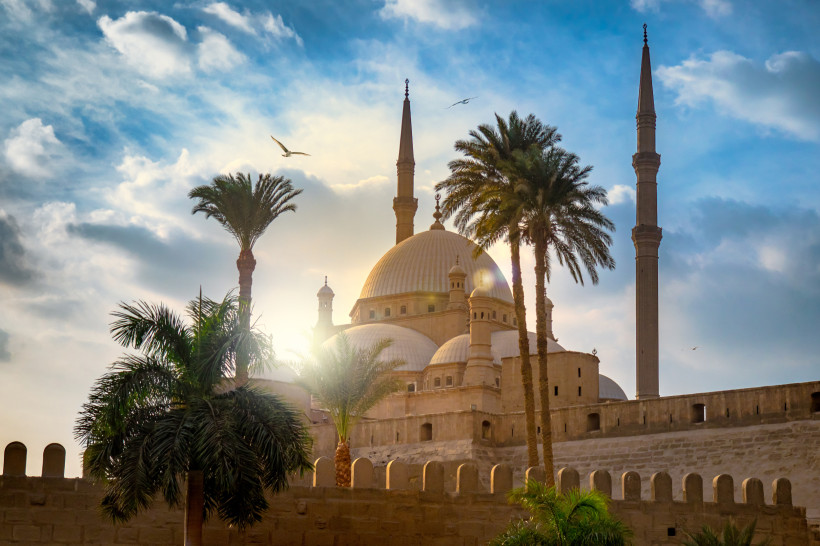 Ägypten Prächtige Moschee mit großer Kuppel und zwei Minaretten, umgeben von hohen Palmen. Die Sonne scheint durch die Wolken und erzeugt ein warmes Lichtspiel hinter dem Gebäude.