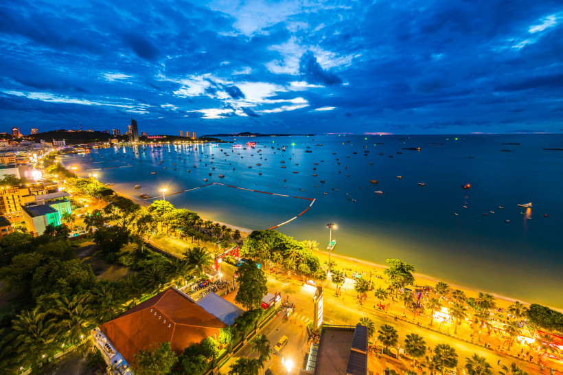 Thailand Pattaya Blick über Pattaya bei Nacht: Die Küste ist hell erleuchtet, zahlreiche Boote liegen in der Bucht. Entlang der Strandpromenade reihen sich Hotels, Palmen und Straßenlichter. Der Himmel ist teils bewölkt, aber noch blau