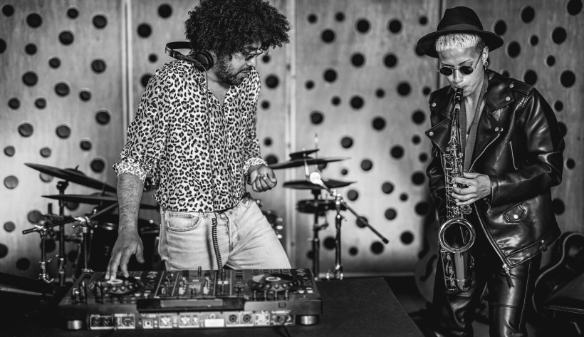Südafrika Schwarz-weiß-Foto eines Musikduos: Links ein DJ mit Leopardenhemd an einem Mischpult, rechts ein Saxophonist mit Sonnenbrille, Hut und Lederjacke beim Spielen des Saxophons. Im Hintergrund Schlagzeug-Setup.
