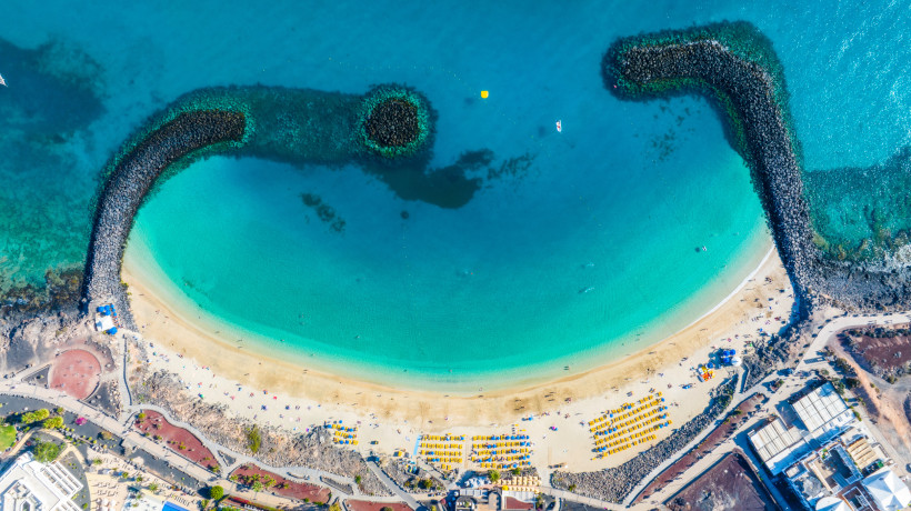 Luftaufnahme der Playa Flamingo auf Lanzarote mit geschützter Bucht, türkisfarbenem Wasser, feinem Sandstrand und Wellenbrechern