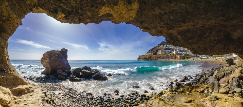 Blick von einer Felsüberhang-Höhle auf die Bucht von Playa del Cura mit Brandung, Kiesstrand und Gebäuden am Hang