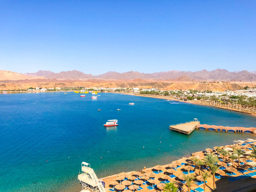 Ägypten Blick auf eine weitläufige Badebucht mit türkisblauem Wasser, gesäumt von Palmen und Sonnenschirmen. Am Ufer führt eine Promenade mit Steg ins Meer. Mehrere Ausflugsboote und Yachten liegen auf dem Wasser. Im Hintergrund sind Wüstenberge und eine flache S