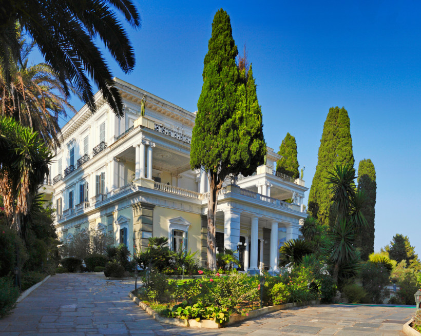 Der Achilleion-Palast auf Korfu, ein weißes klassizistisches Gebäude mit Säulen, Balkonen und Statuen auf dem Dach. Umgeben von hohen Zypressen, Palmen und Blumenbeeten vermittelt der Palast ein majestätisches Flair unter strahlend blauem Himmel.