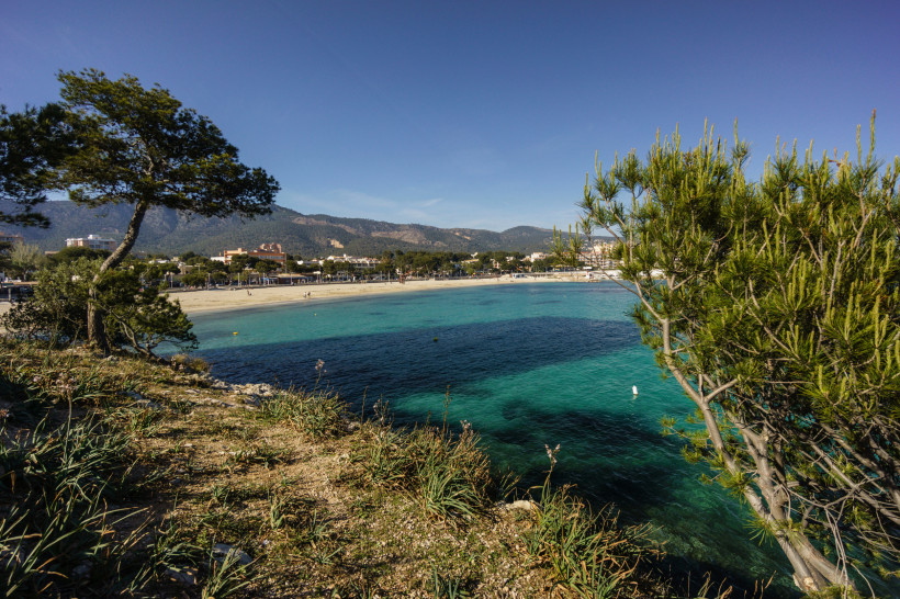 Bucht Es Carregador mit Sandstrand und türkisfarbenem Meer, im Hintergrund Bebauung und Berge