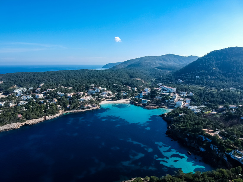 Ibiza - Portinatx Luftaufnahme eines kleinen Ferienortes im Norden Ibizas. Der Ort liegt zwischen bewaldeten Hügeln am türkisblauen Meer mit Stränden, Hotels und Ferienhäusern.