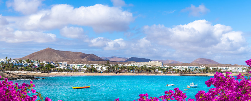 Panoramablick auf Costa Teguise auf Lanzarote mit hellem Sandstrand, türkisblauem Meer, weißen Häusern und Bergen im Hintergrund.