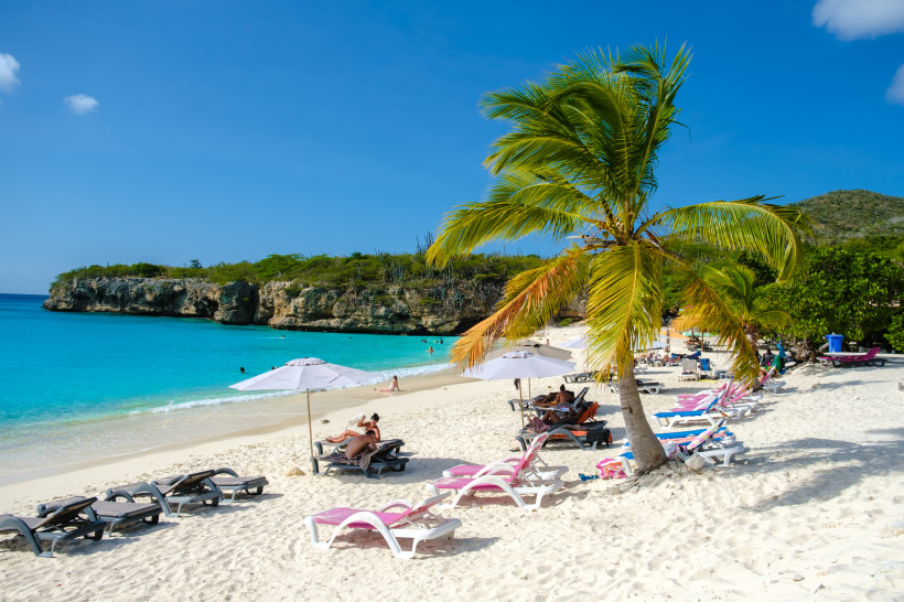 Grote Knip Strand – Karibiktraum auf Curaçao mit Palmen und türkisblauem Wasser Grote Knip Strand auf Curaçao – Sandstrand mit Palmen, Sonnenliegen, türkisblauem Meer und Felsküste im Hintergrund