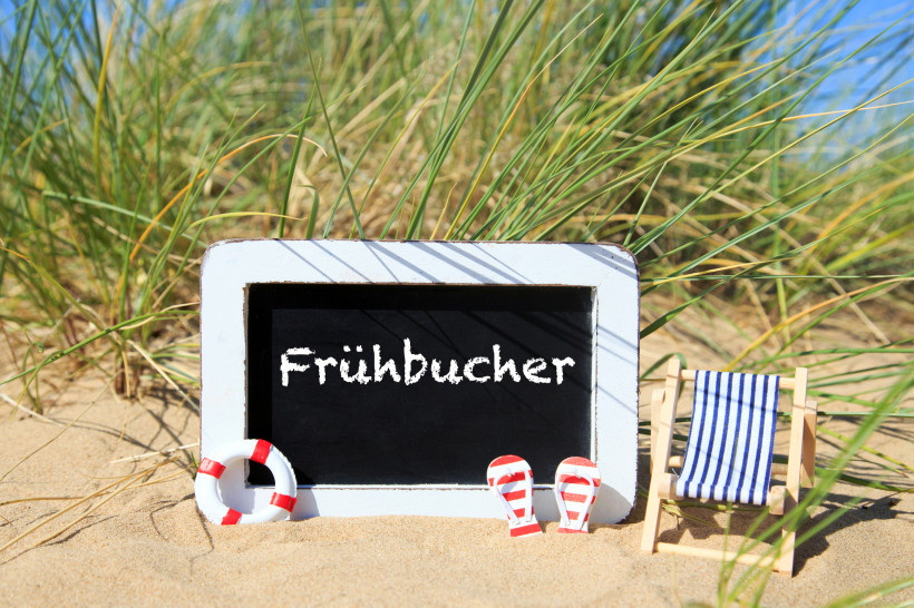 Kleine Tafel mit der Aufschrift „Frühbucher“ steht im Sand zwischen Dünengras, daneben ein Miniatur-Liegestuhl, Flip-Flops und ein Rettungsring.