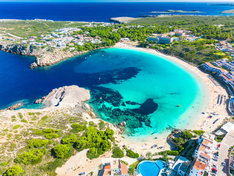 Luftaufnahme einer sichelförmigen Bucht mit türkisblauem, glasklarem Wasser auf Menorca, Spanien. Der weiße Sandstrand ist von Sonnenschirmen gesäumt und von Hotels sowie Ferienhäusern eingerahmt. Im Vordergrund ein Felsen und ein großes rundes Hotelpool,