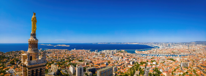 Marseille Weitwinkliges Stadtpanorama einer südfranzösischen Küstenmetropole. Links im Vordergrund ragt ein Turm mit einer goldenen Marienstatue auf. Darunter breitet sich die Stadt mit vielen orangefarbenen Ziegeldächern und modernen Hochhäusern aus. M