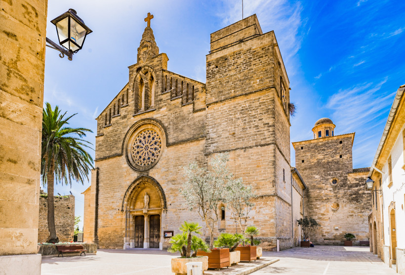 Blick auf die Kirche Sant Jaume in der Altstadt von Alcudia mit gotischer Fassade, angrenzender Stadtmauer und kleinem Platz im historischen Ortskern